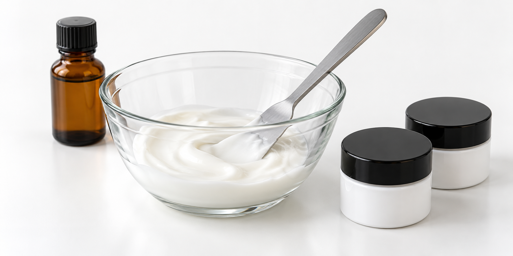 A glass bowl of white lotion with a mixing spatula beside an amber bottle and two closed sample jars.